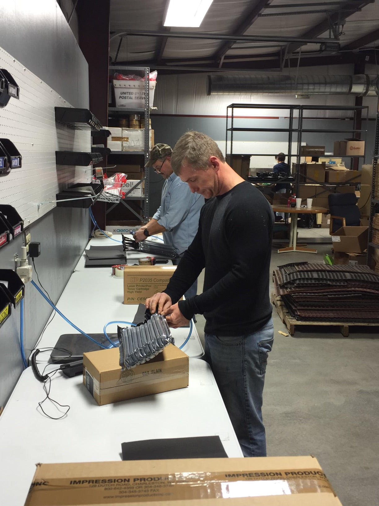 Impression Products team members working in a warehouse, packaging and inspecting printer components at a workstation.