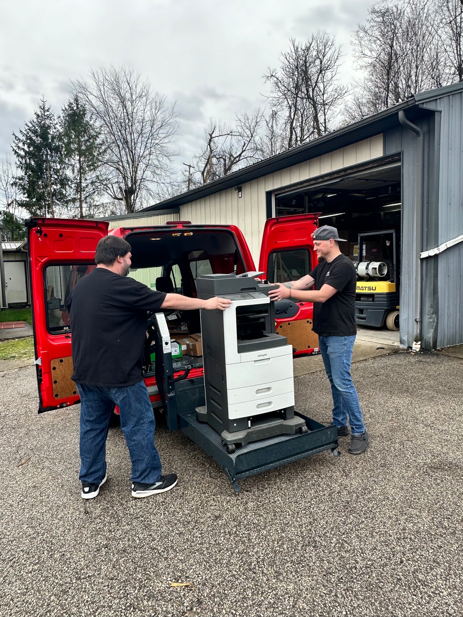 Two technicians loading a printer into a red service van, highlighting logistics for delivering cartridges and equipment.