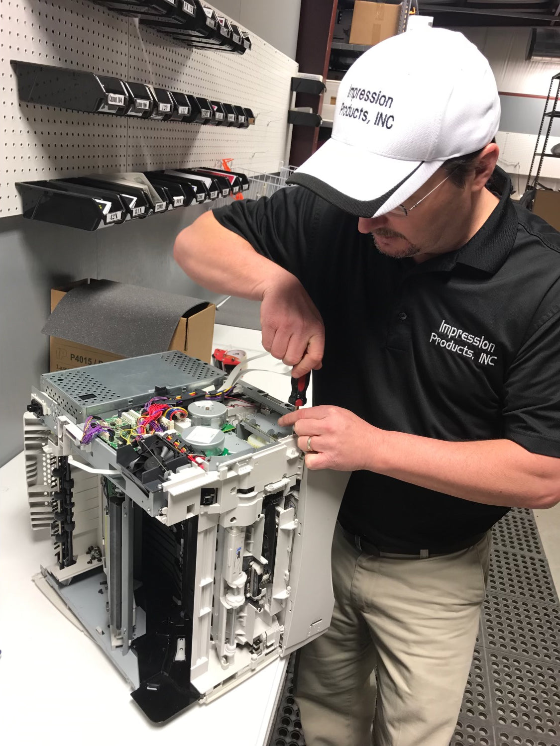 Impression Products technician repairing a printer at a workstation, wearing branded apparel and using tools for maintenance.