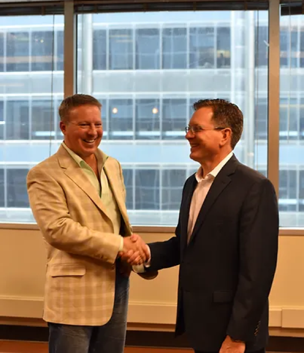 Two businessmen shaking hands in front of a large office window, with Eric Smith on the left wearing a beige blazer.