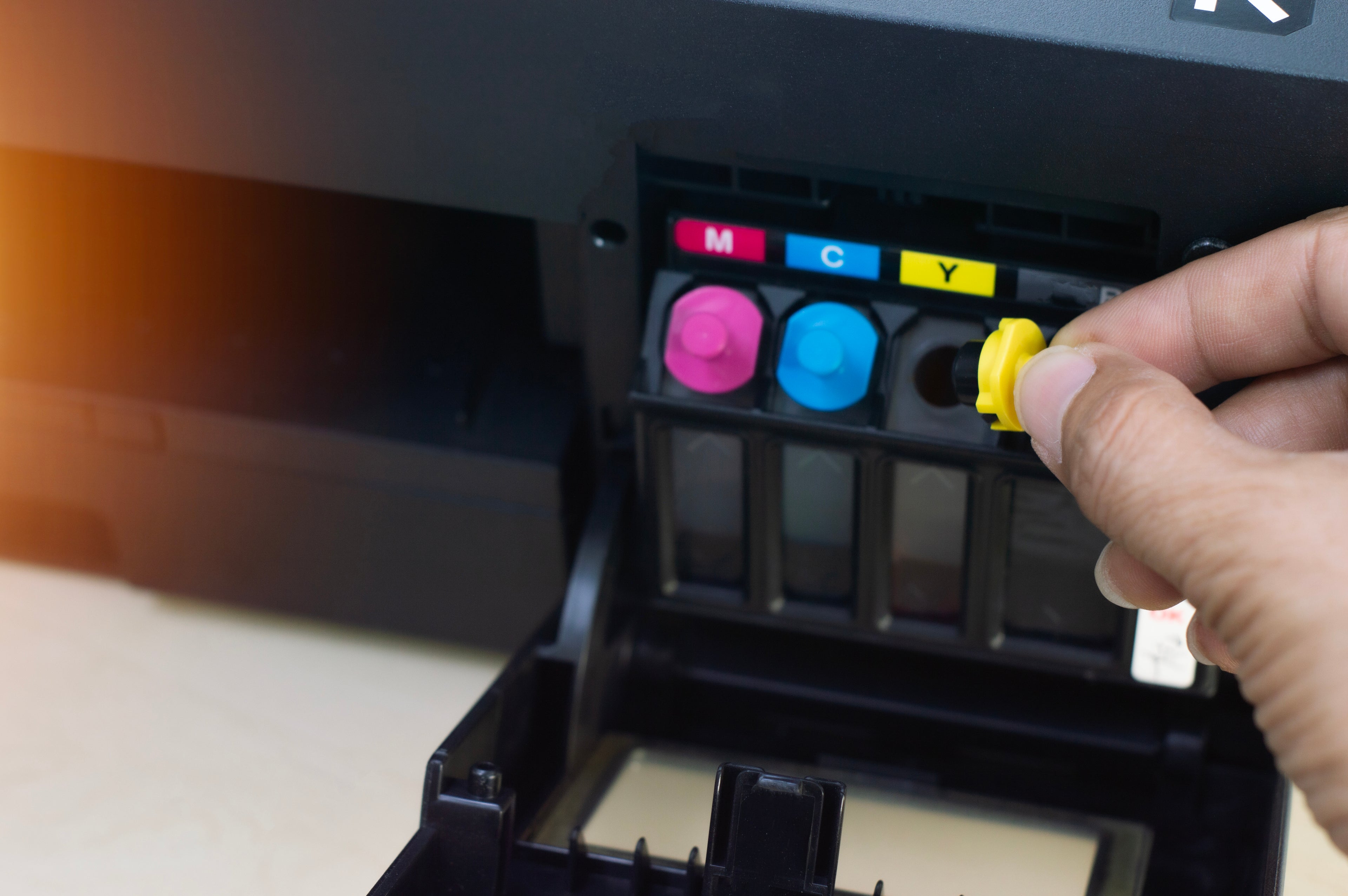 Close-up of a person replacing Remanufactured Ink Cartridges in a color printer, showing cyan, magenta, and yellow ink tanks.